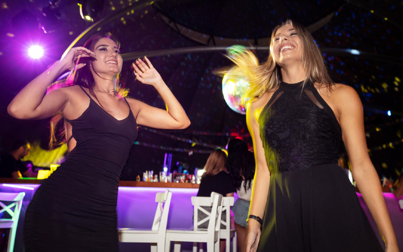 Dos mujeres bailando en una fiesta latina privada de cumpleaños en Madrid, con luces de discoteca y barra al fondo